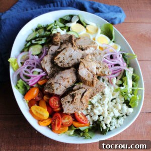 Large serving bowl filled with salad topped with blue cheese, tomatoes, pickled red onions, hard boiled eggs, cucumbers and sliced steak with chimichurri sauce on top, ready to be served.