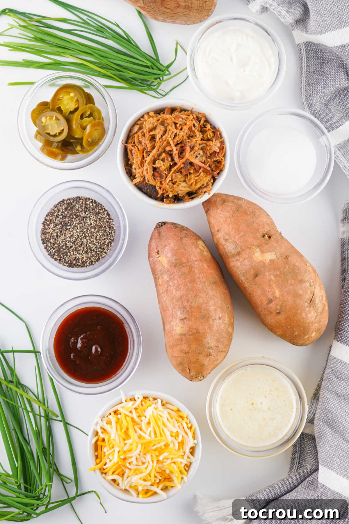 An array of fresh ingredients for stuffed sweet potatoes, including whole sweet potatoes, various cheeses, salt, pepper, pre-cooked pulled pork, BBQ sauce, sour cream, fresh chives, butter, and sliced jalapenos.
