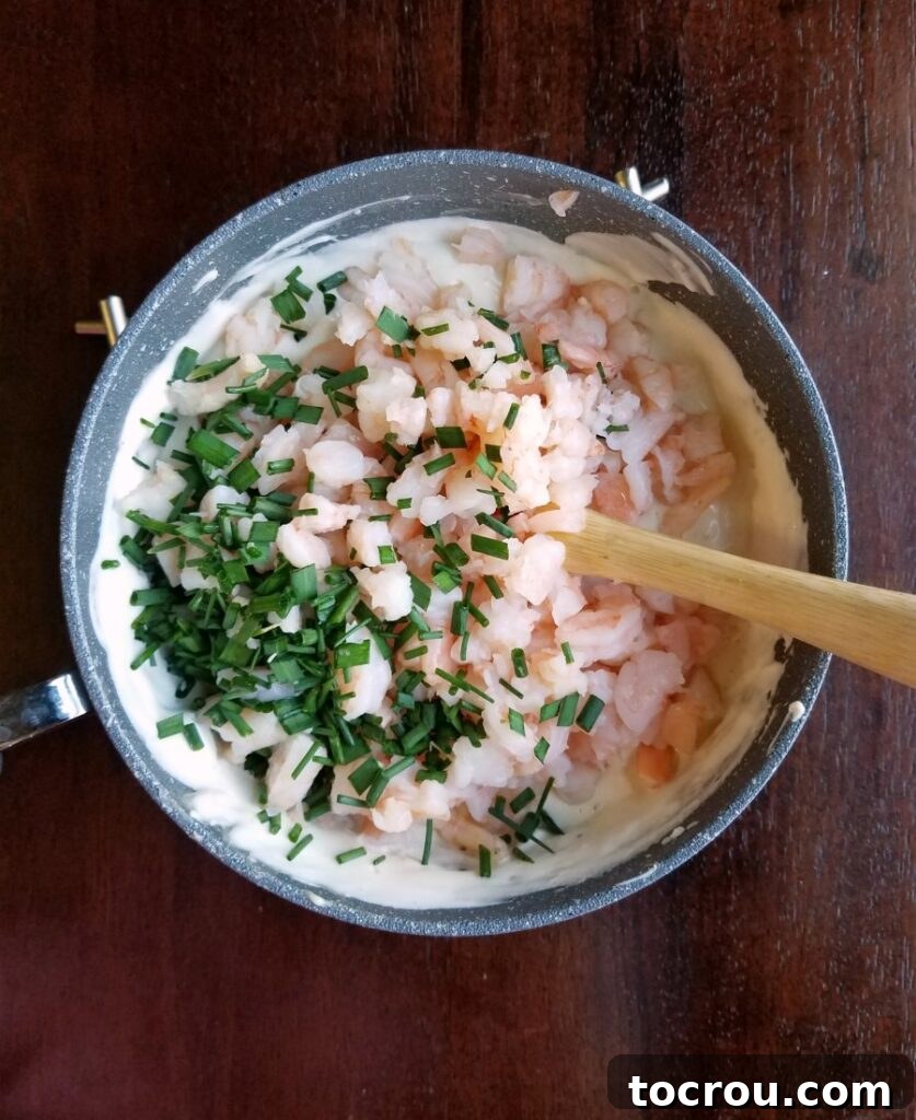 Saucepan with a creamy mixture, chopped shrimp, and fresh chives, ready to be combined into the dip.