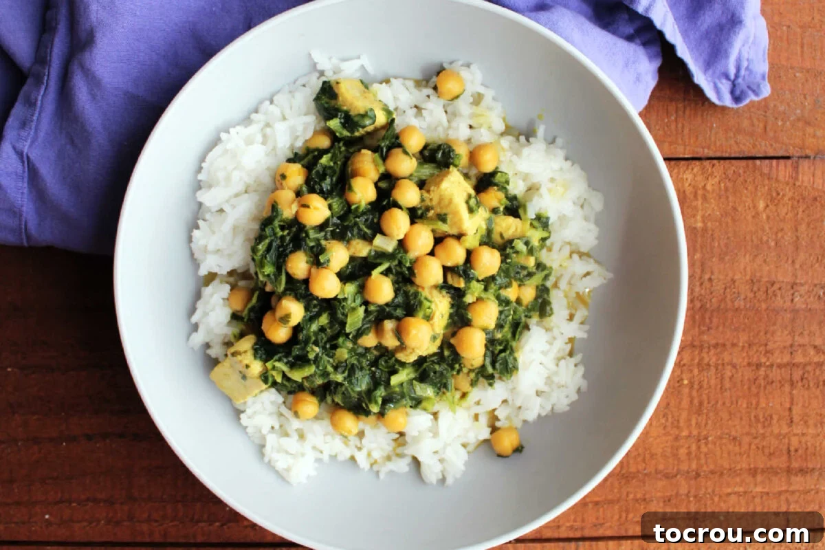 Close up of a bowl of coconut milk based chicken and chickpea curry with yellow broth and bits of spinach inside served over white jasmine rice, ready to eat. 