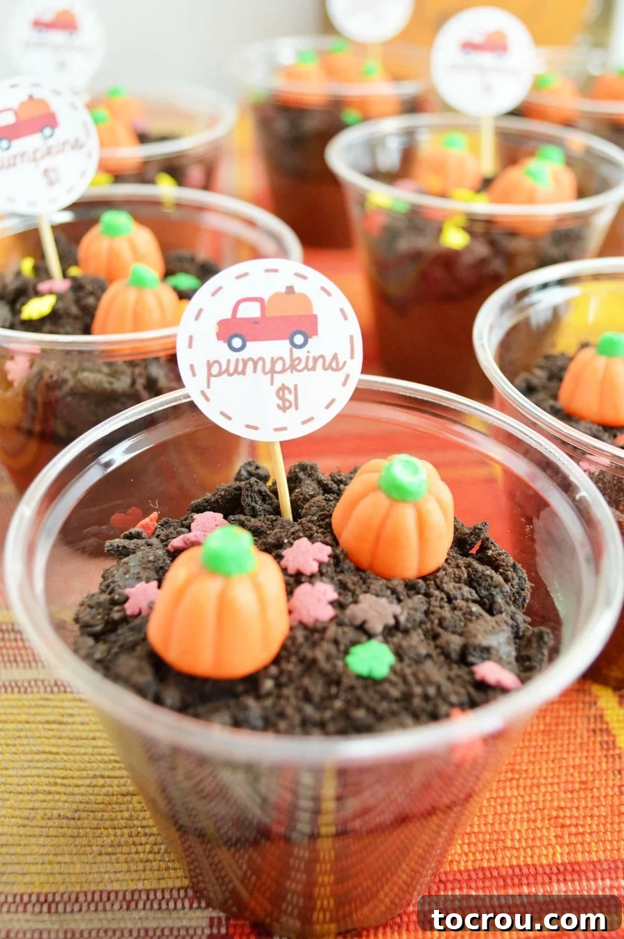 Small clear plastic cup filled with chocolate pudding and chocolate cookie crumbs to look like dirt topped with candy pumpkins and a pick with a red truck and pumpkin on it. 