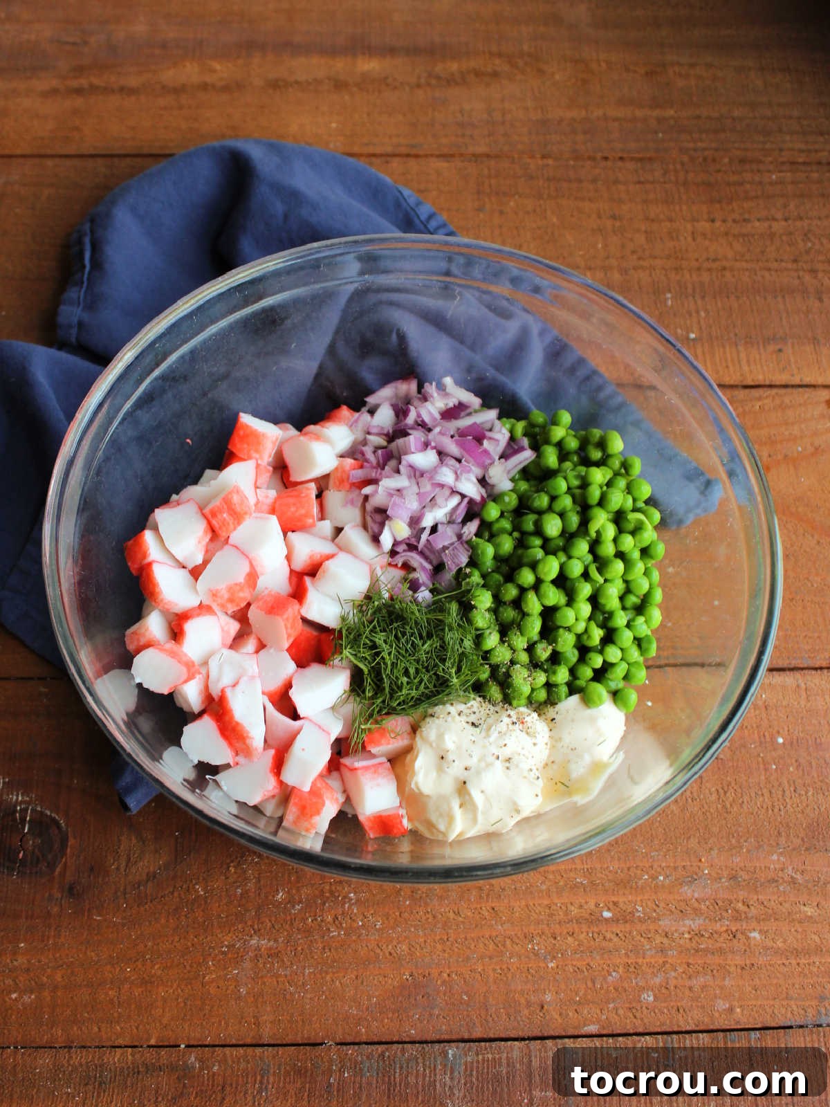 Large glass mixing bowl with mayonnaise, salt, pepper, fresh dill, peas, diced red onion, and chopped imitation crab inside.