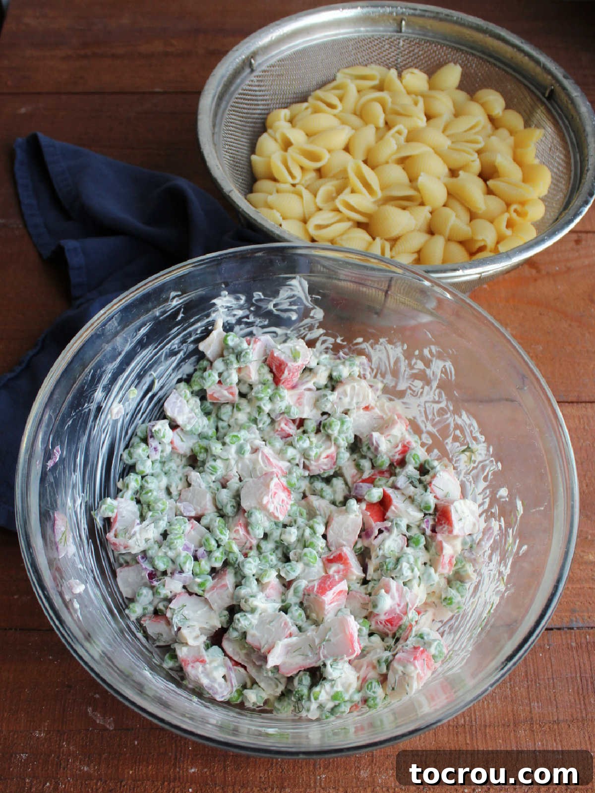 Strainer with cooked and drained shell pasta next to mixing bowl with crab and peas in creamy dressing, ready to be mixed together into pasta salad. 
