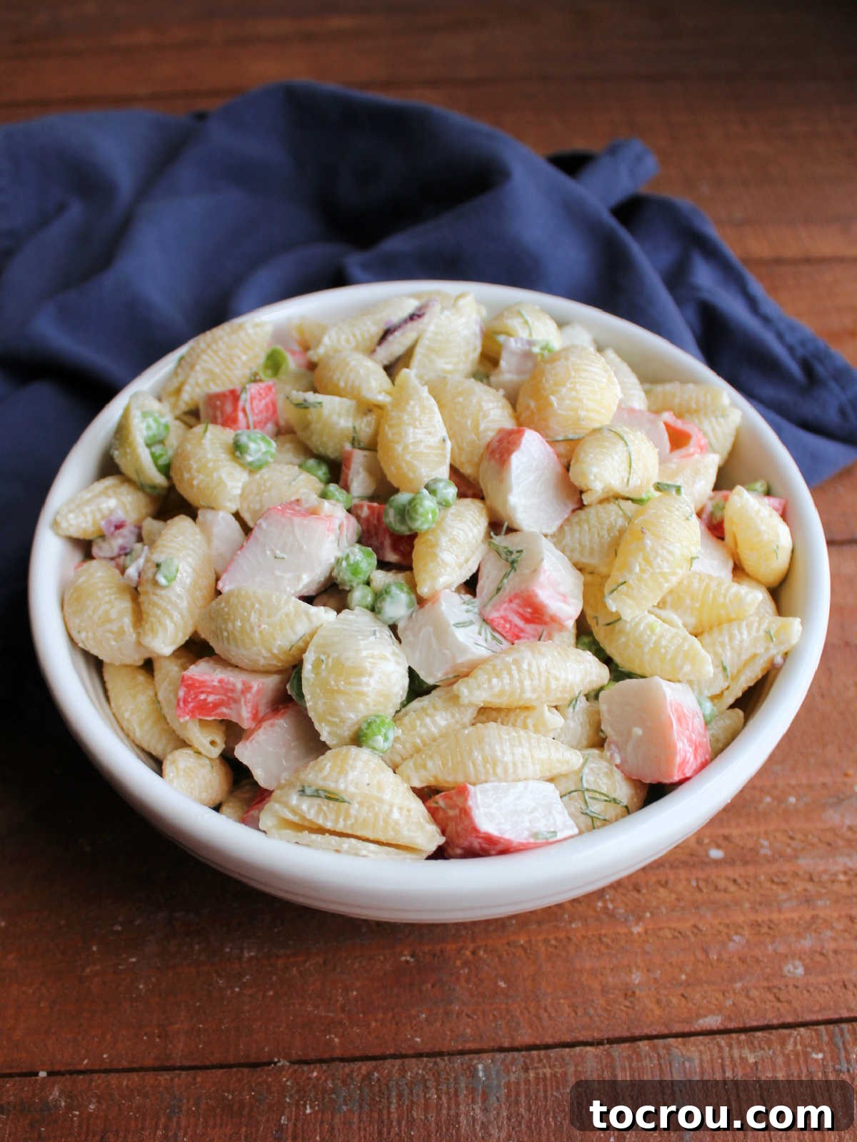 White serving bowl piled high with creamy seafood pasta salad with peas and dill. 