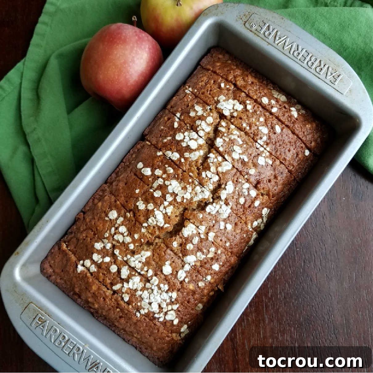 A perfectly sliced loaf of applesauce oatmeal quick bread resting in its loaf pan, garnished with fresh apples, ready to be enjoyed.