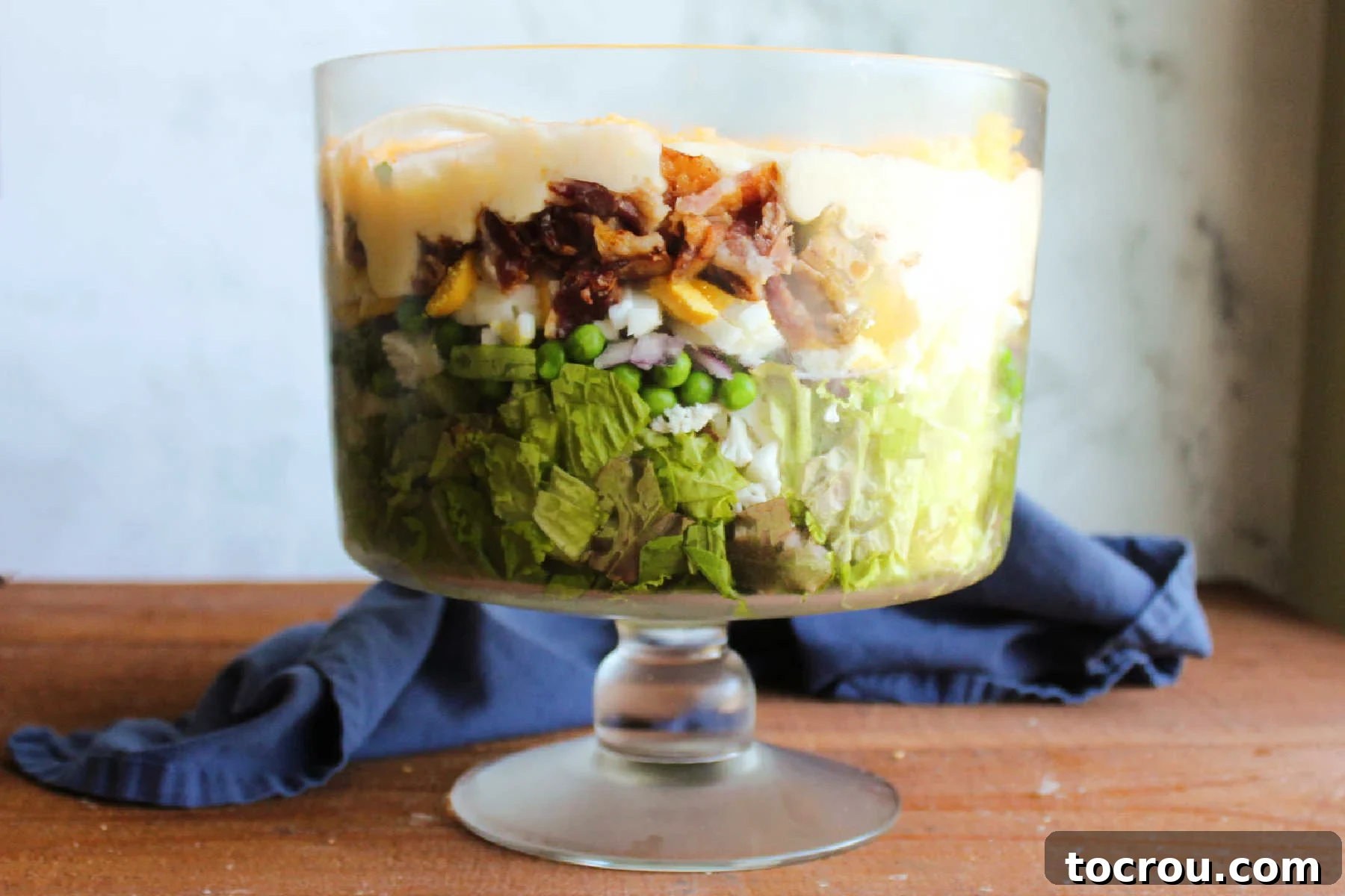 Glass bowl filled with seven layer salad made of lettuce, peas, onion, cauliflower, bacon, eggs, dressing and cheese. 