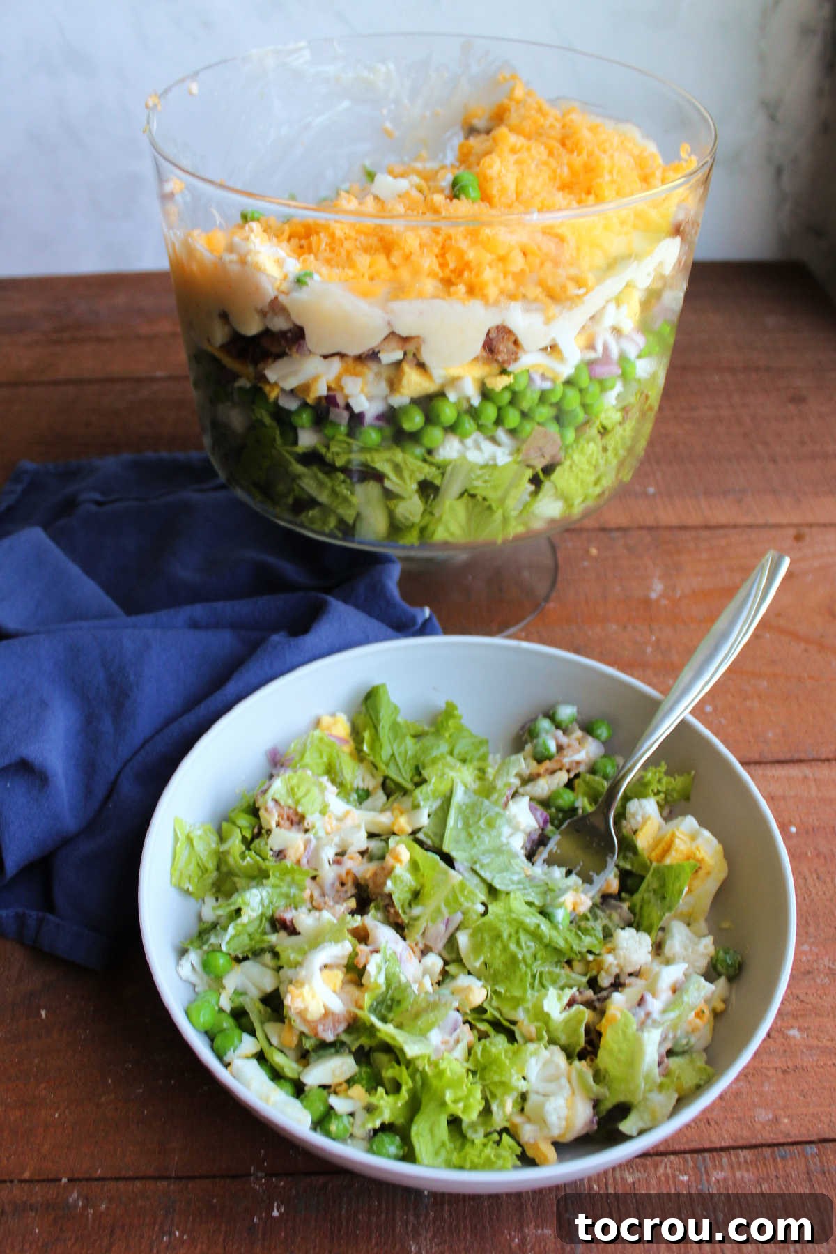 Bowl of salad with creamy dressing and lots of toppings next to trifle bowl with remaining seven layer salad inside. 