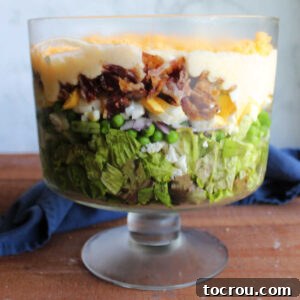 Glass trifle bowl filled with a big layer of lettuce topped with peas, cauliflower, onion, eggs, bacon, and a creamy dressing making a seven layer salad.