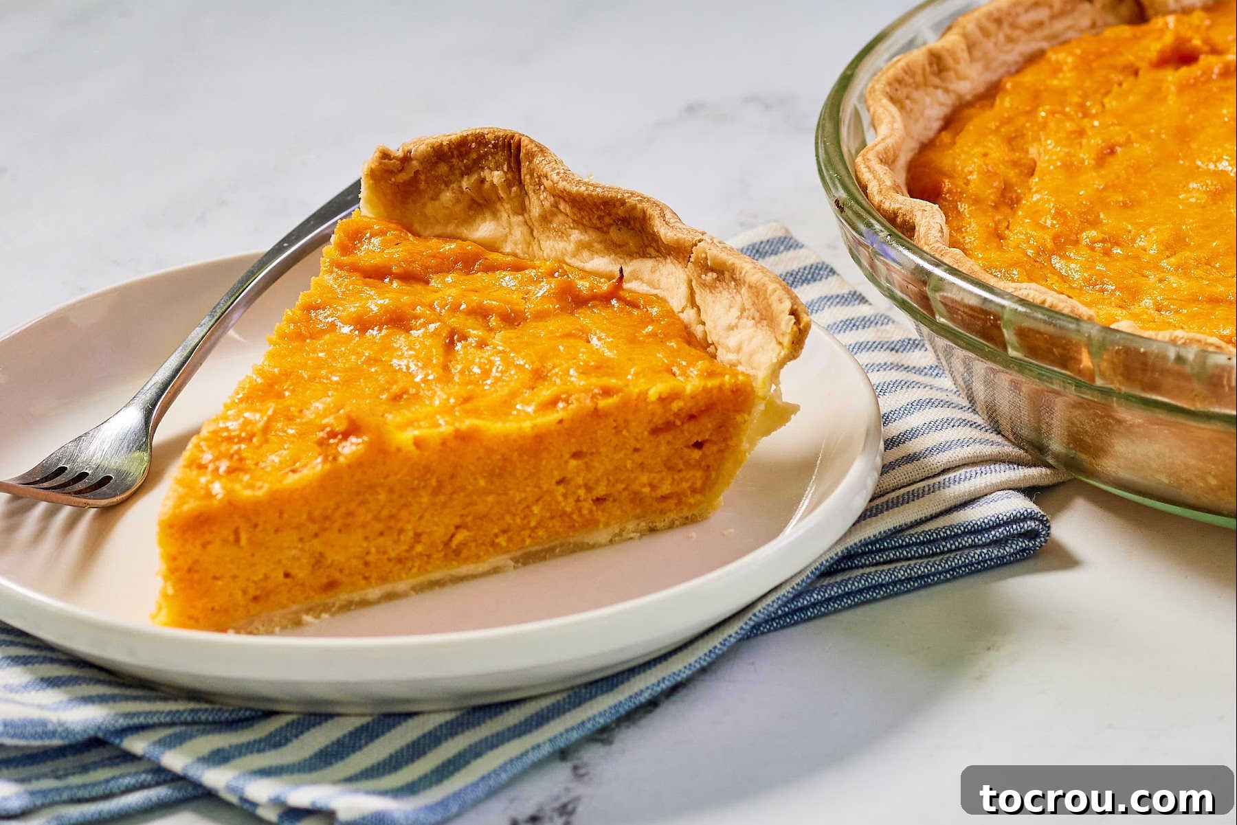 Decadent Sweet Potato Pie with Condensed Milk 2 Slice of sweet potato pie on dessert plate with fork with the remaining pie in the background.