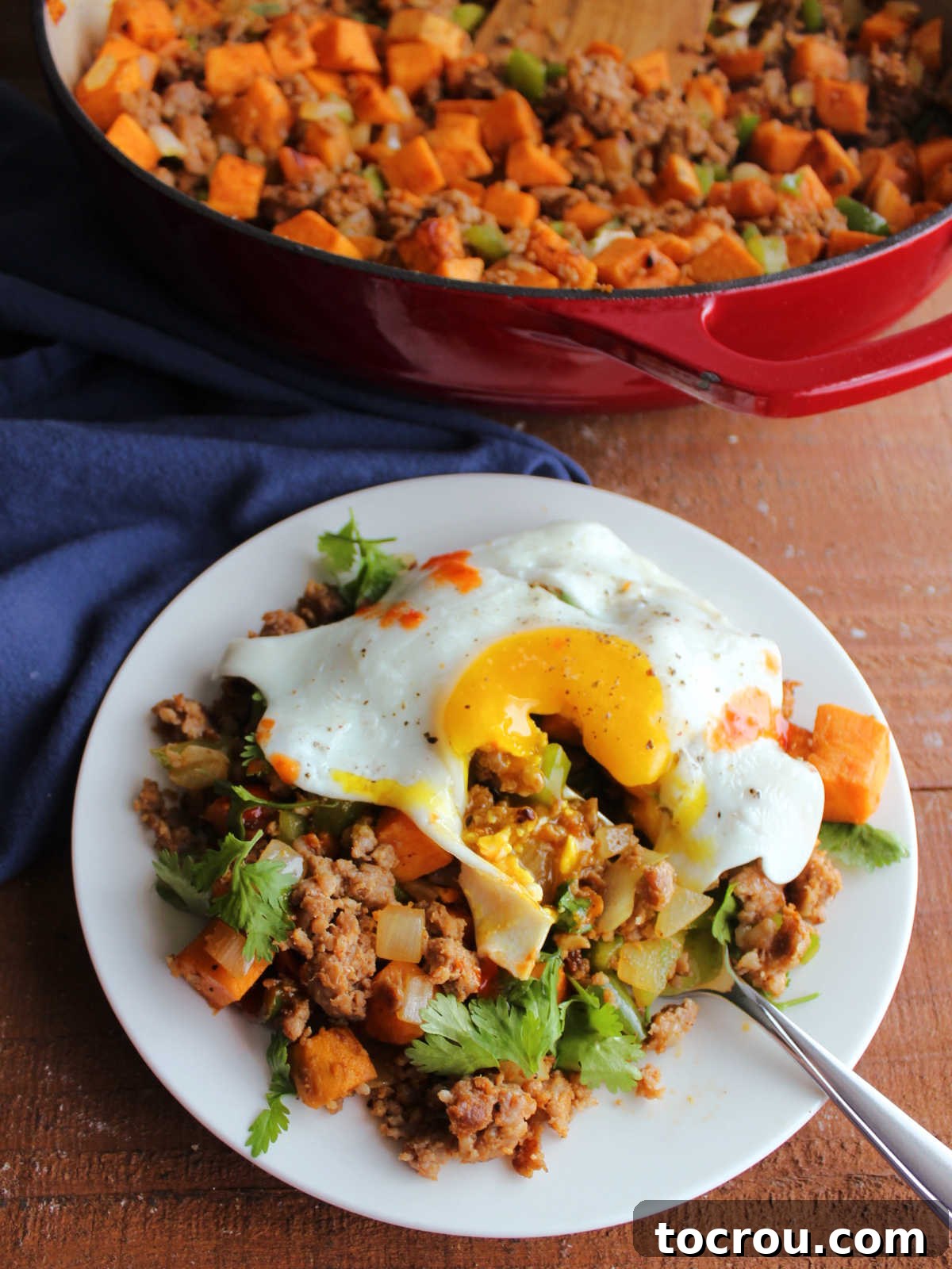 A fork piercing the bright yellow yolk of a fried egg, allowing the rich, runny yolk to flow down onto the flavorful sweet potato chorizo hash below, creating a mouth-watering visual.