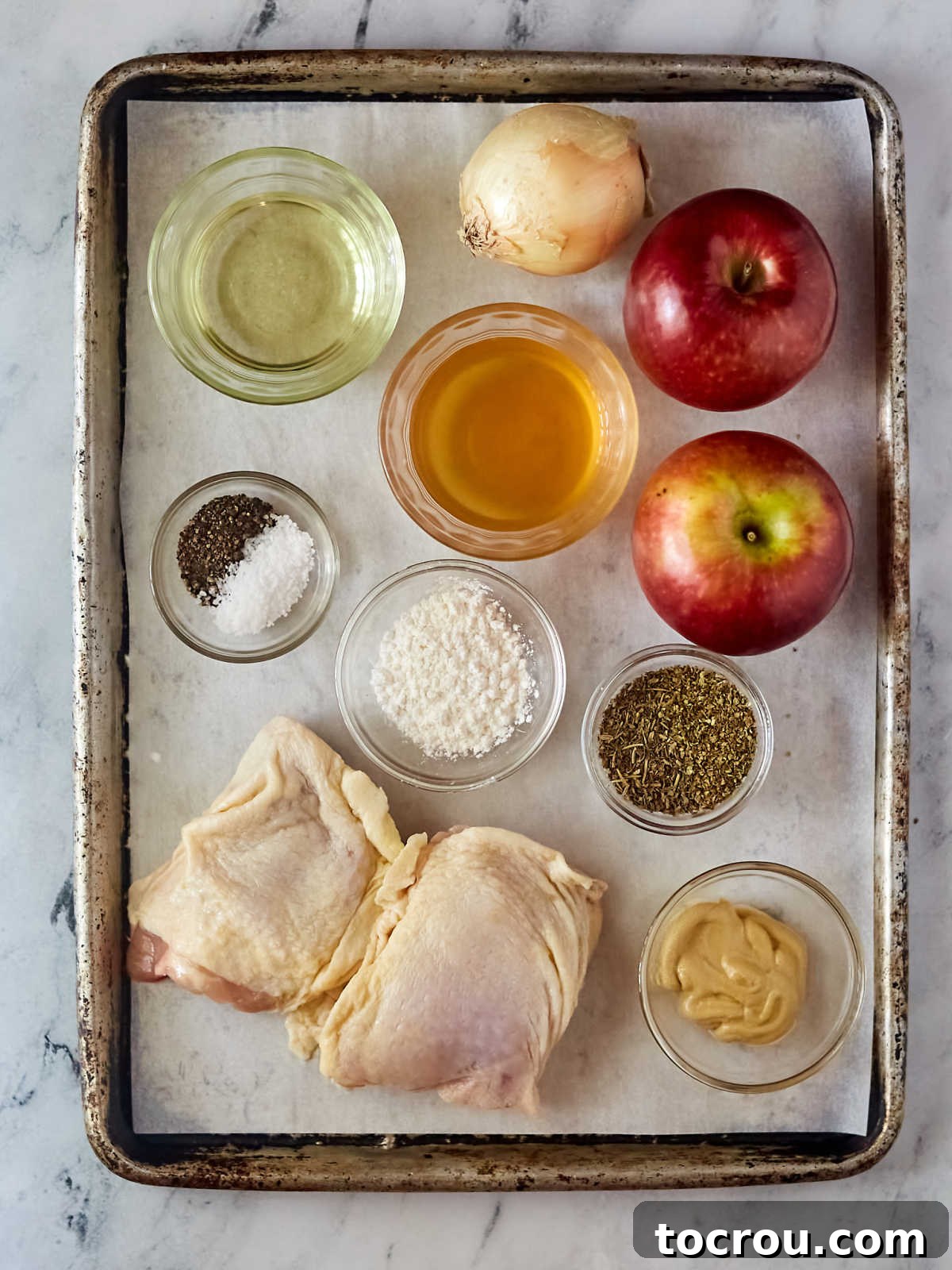 Ingredients including chicken thighs, apples, an onion, apple cider, oil, salt, pepper, mustard, rosemary, and sage ready to be made into skillet apple cider chicken.