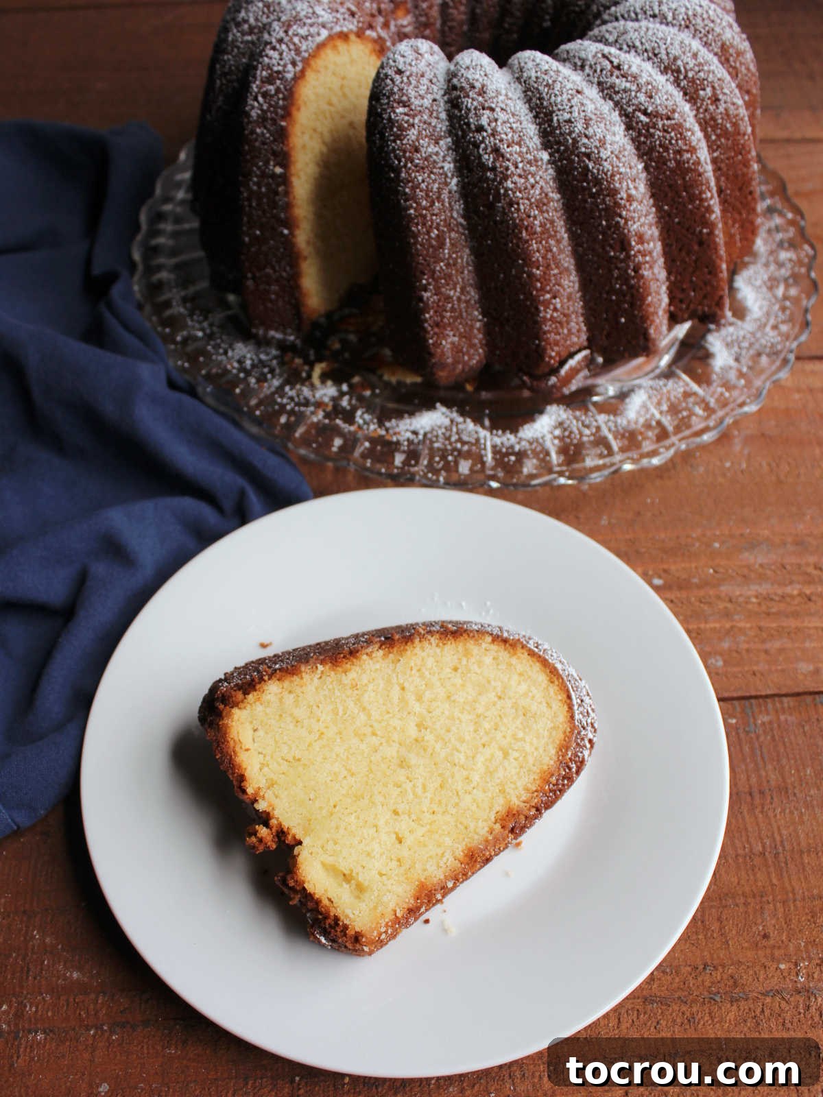 Piece of pound cake with dark crust and creamy center on plate with remaining powder sugar dusted bundt cake in the background.