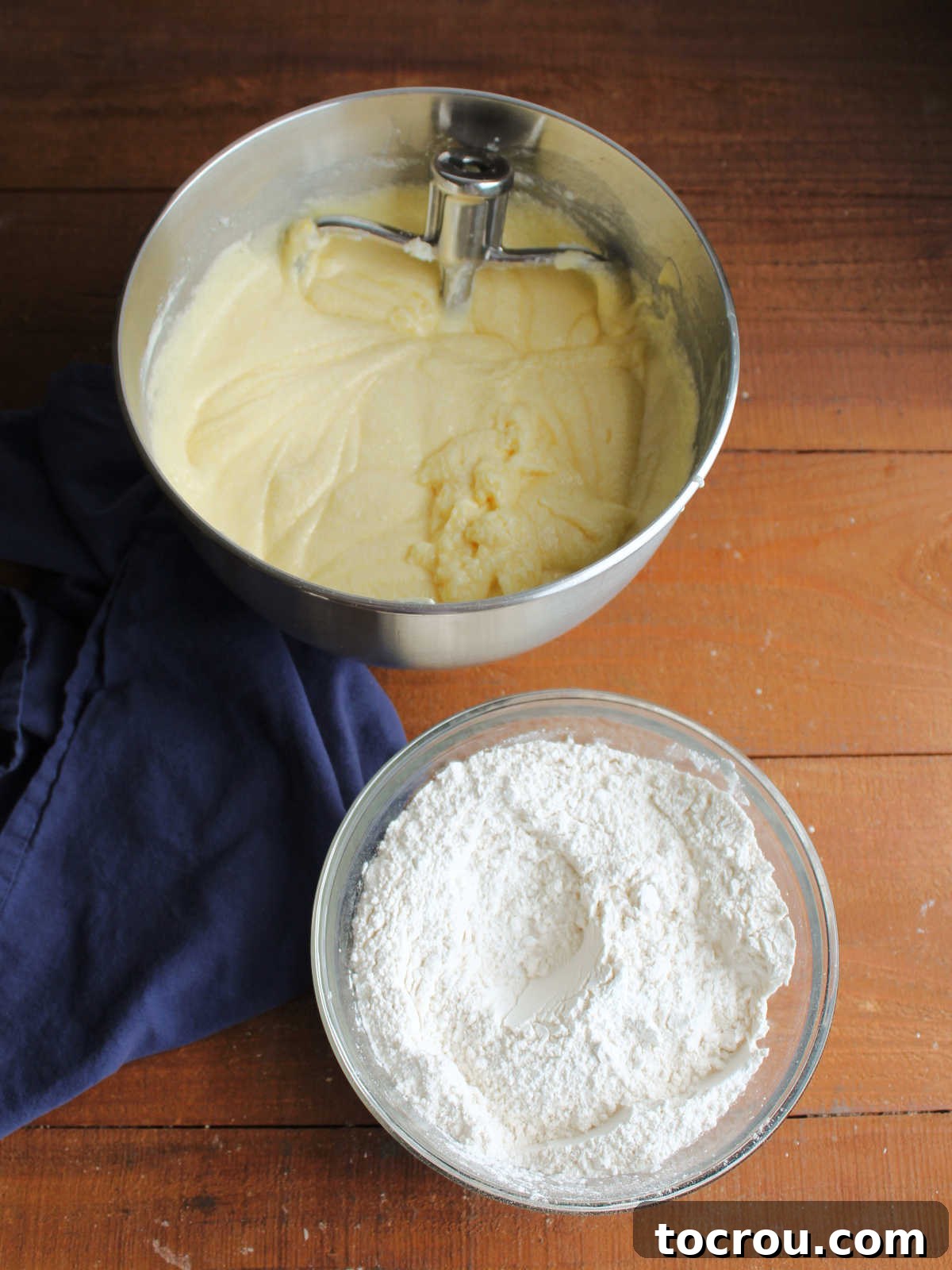 Mixing bowl with wet ingredients mixture including butter, sugar, condensed milk, eggs and flavorings next to mixing bowl with dry ingredient mixture of flour, baking powder and salt, ready to be combined to make cake batter.