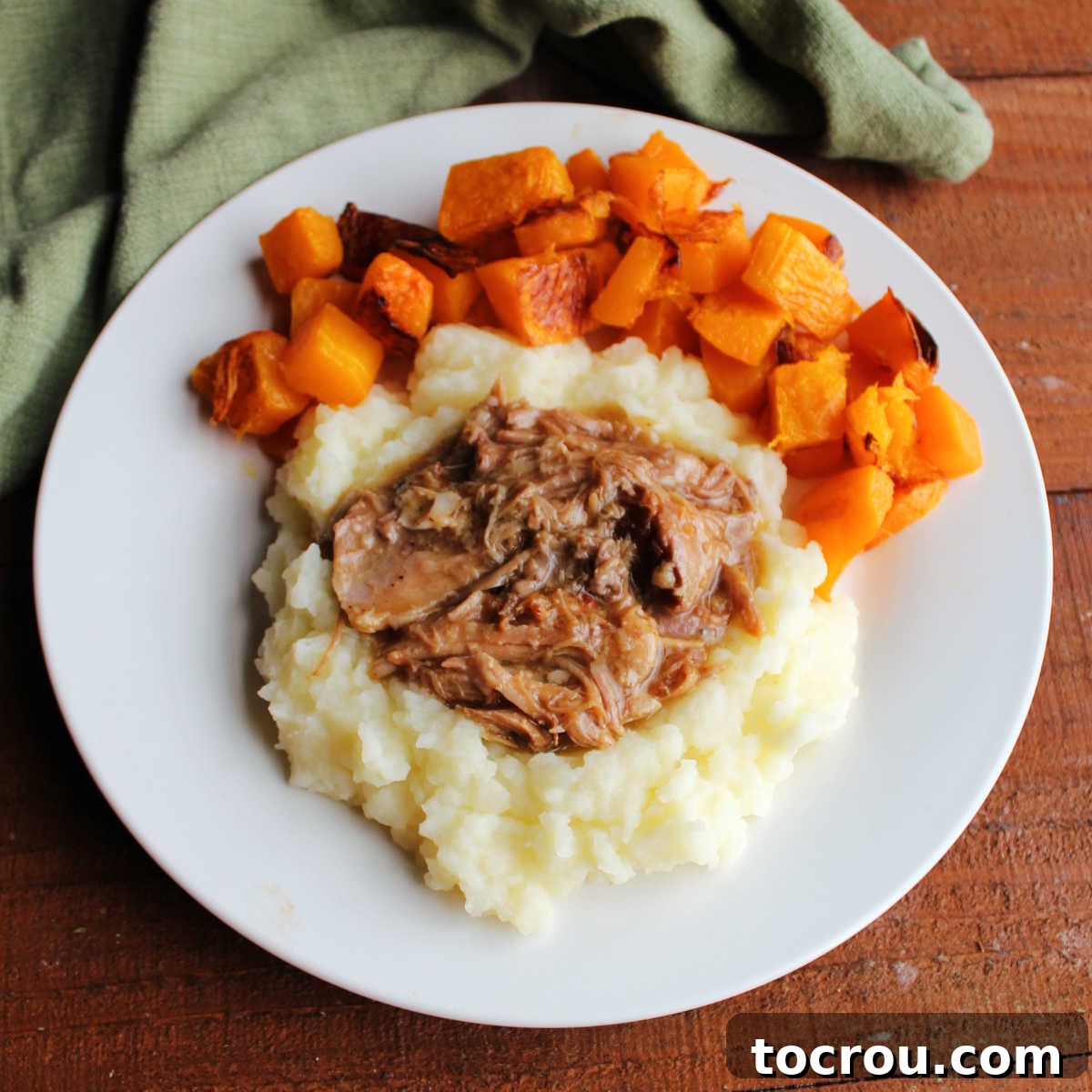 Slow cooked apple cider pulled pork served on mashed potatoes with a side of roasted butternut squash, showcasing the rich, tender meat and savory gravy.