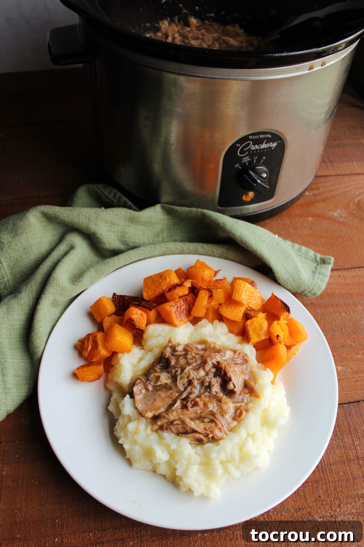 A perfectly plated dinner featuring apple cider pulled pork atop mashed potatoes, accompanied by roasted butternut squash, with the remaining pork mixture visible in a crockpot in the background.