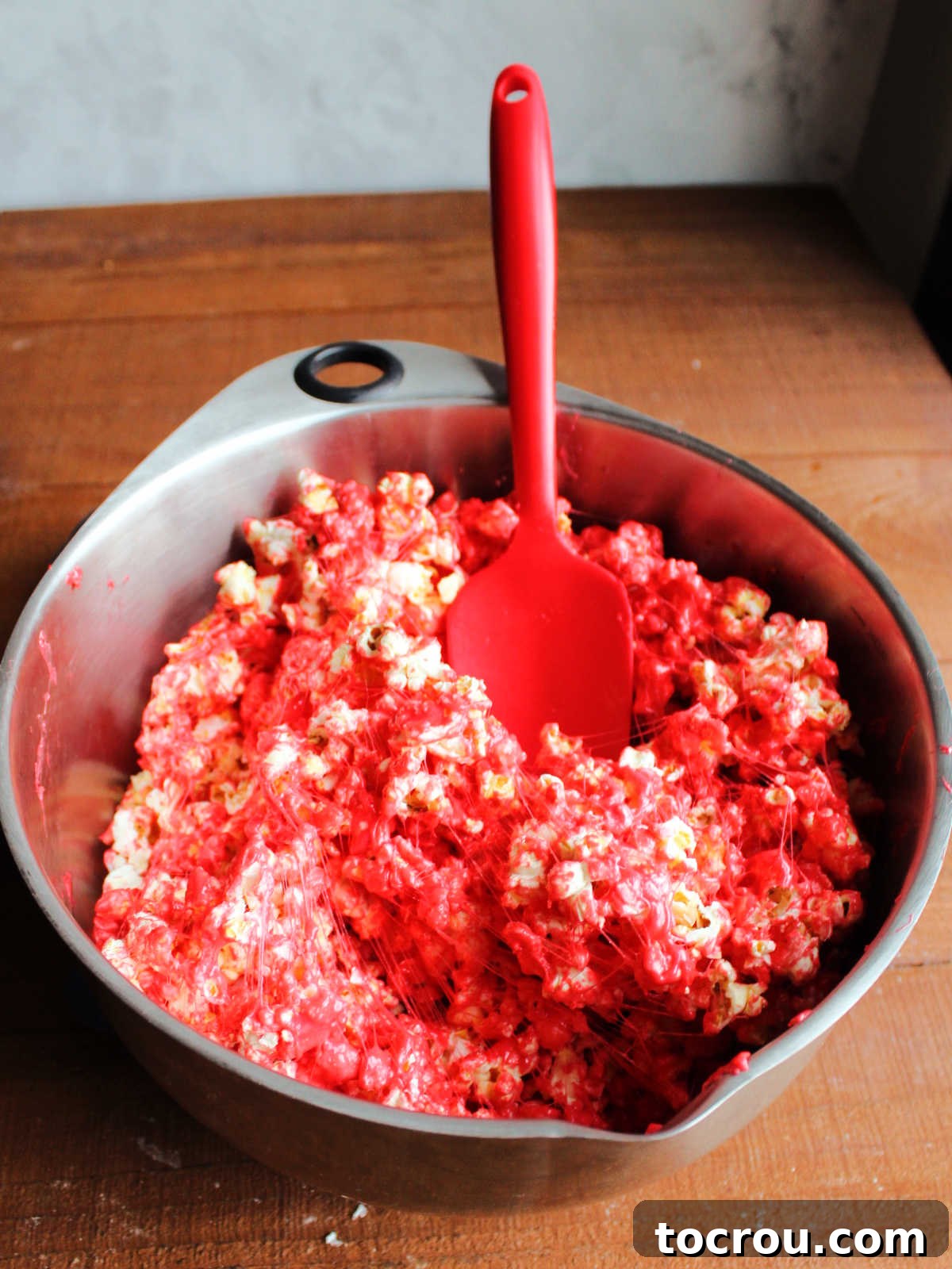 Large mixing bowl filled with popcorn coated in a red Jell-O and marshmallow mixture, ready to be formed into popcorn balls.