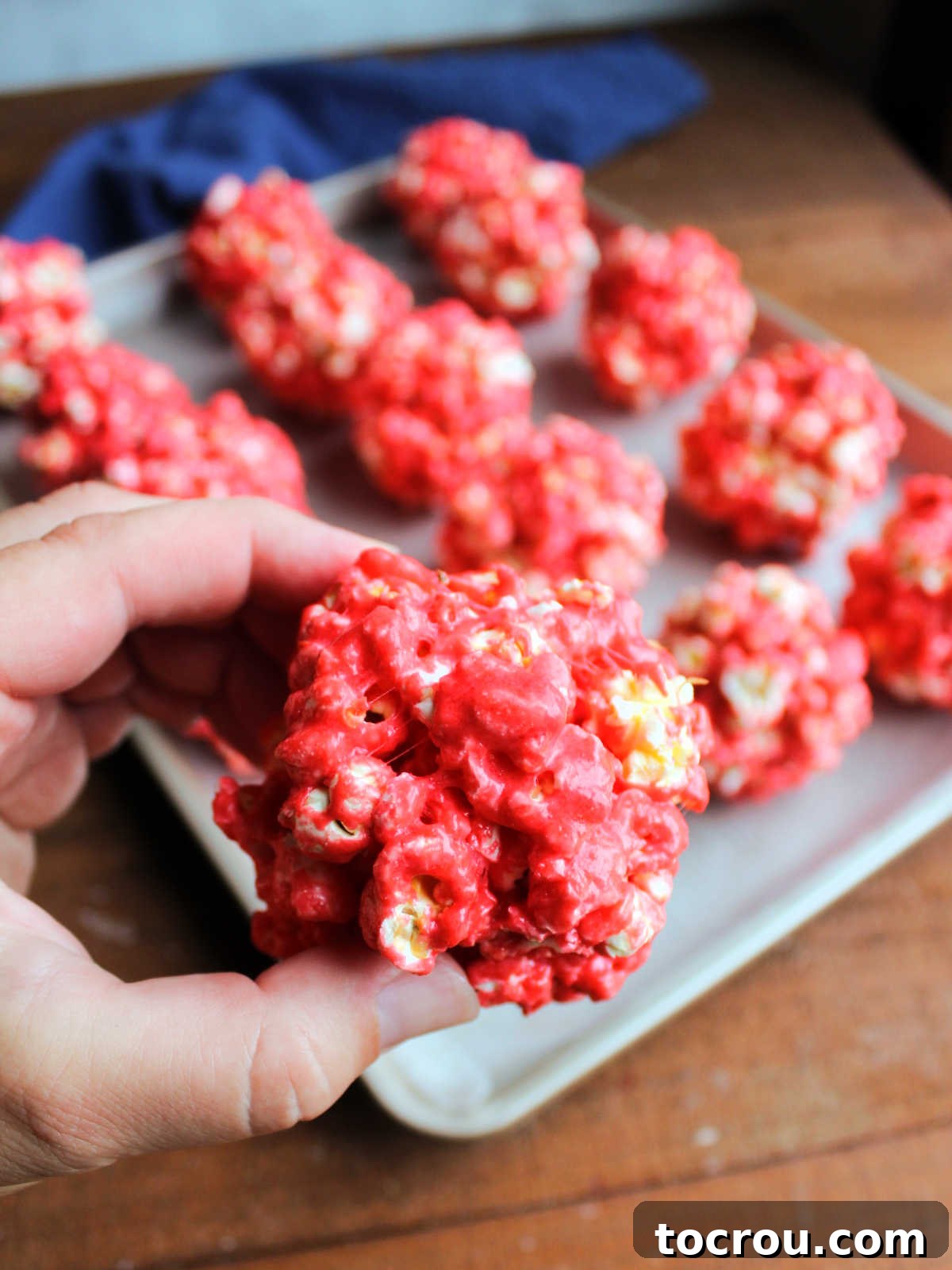 Hand holding a freshly made red Jell-O popcorn ball, with more red popcorn balls resting in the background.