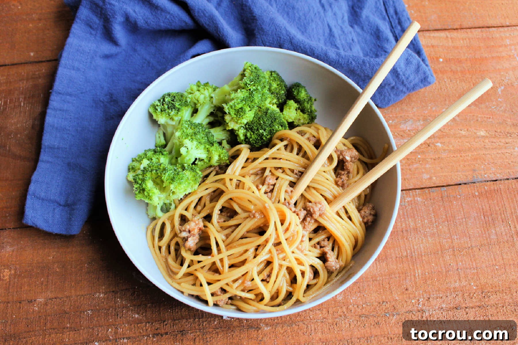 Shallow bowl with Mongolian ground pork and noodles, garnished with sesame seeds and served with steamed broccoli and chopsticks.
