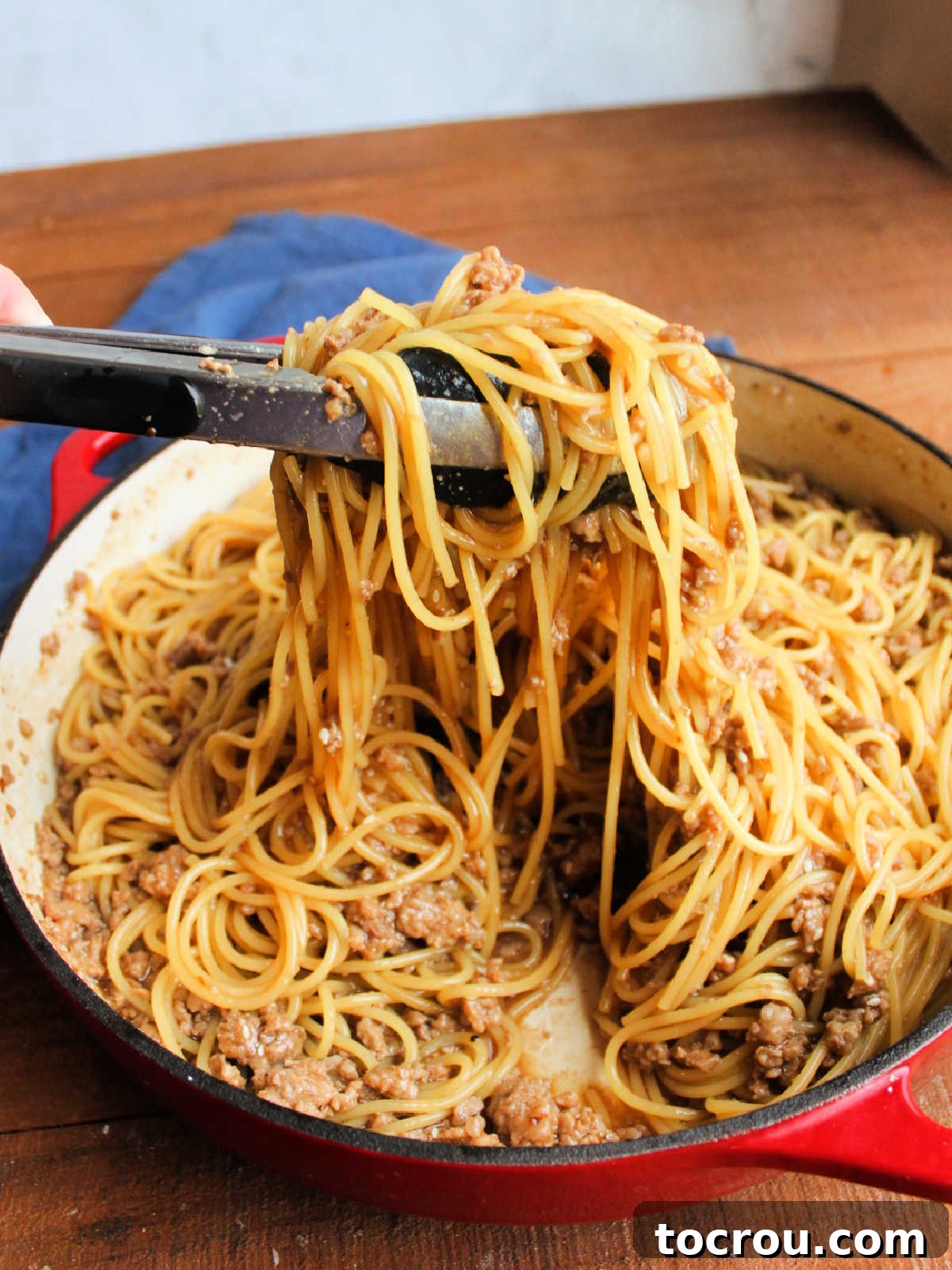 Tongs lifting a generous serving of spaghetti noodles with Mongolian ground pork and savory sauce, ready to be plated.