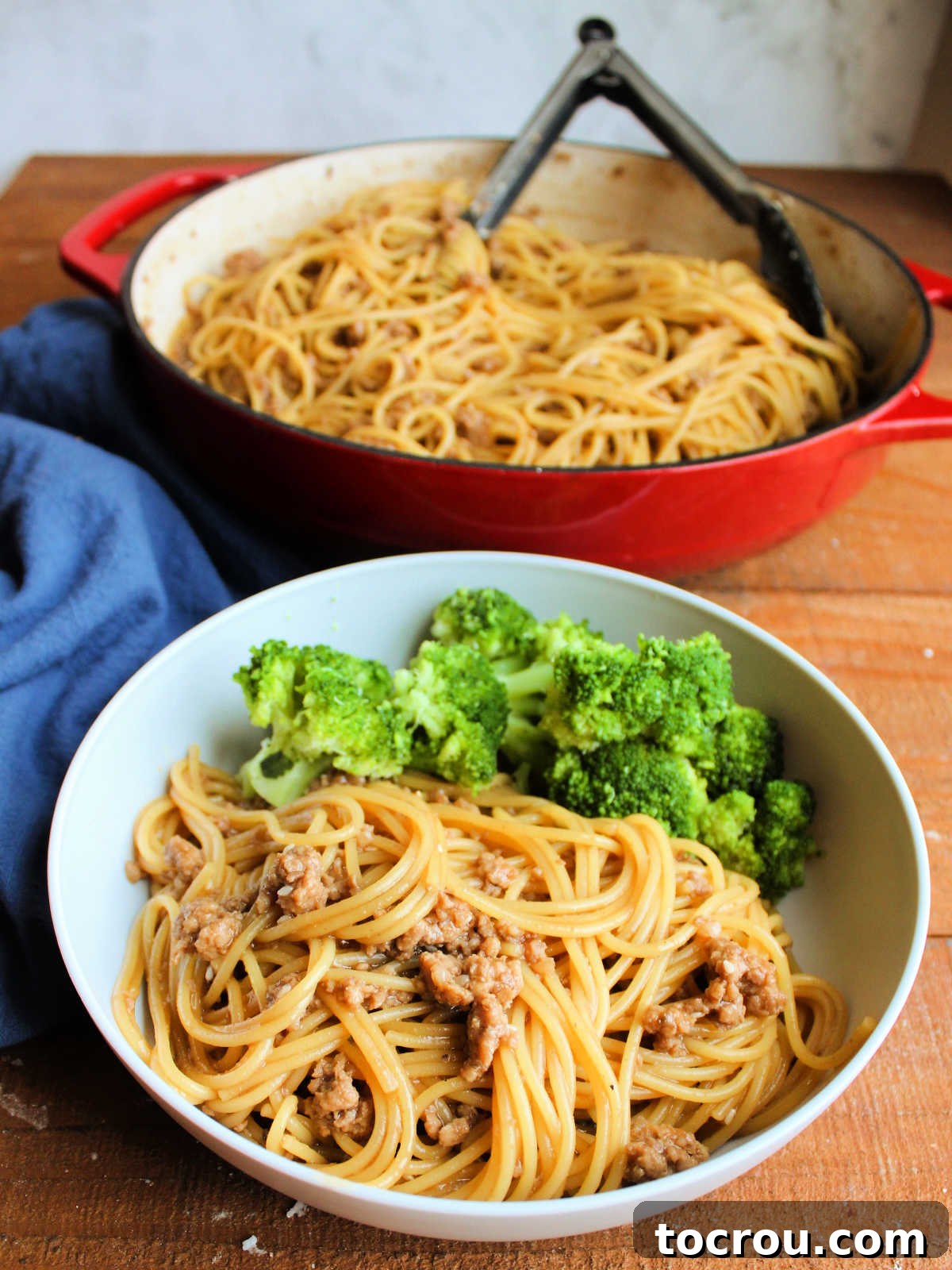A bowl filled with a serving of Mongolian ground pork noodles and steamed broccoli, with the large pan of remaining noodles blurred in the background, ready for more servings.