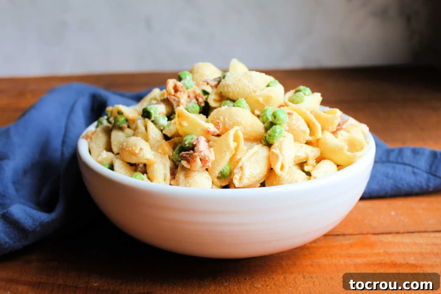 Serving bowl piled high with creamy macaroni salad with bright green peas, crispy bacon bits, and shredded cheddar cheese, ready to be enjoyed.