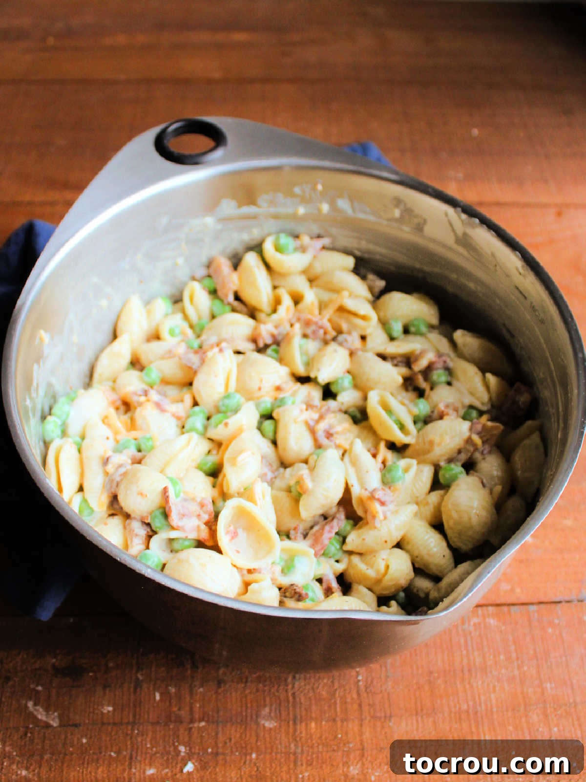 Mixing bowl filled with shell pasta coated in creamy dressing with peas, bacon, and cheese after it has all been stirred together, showing the completed mixture.