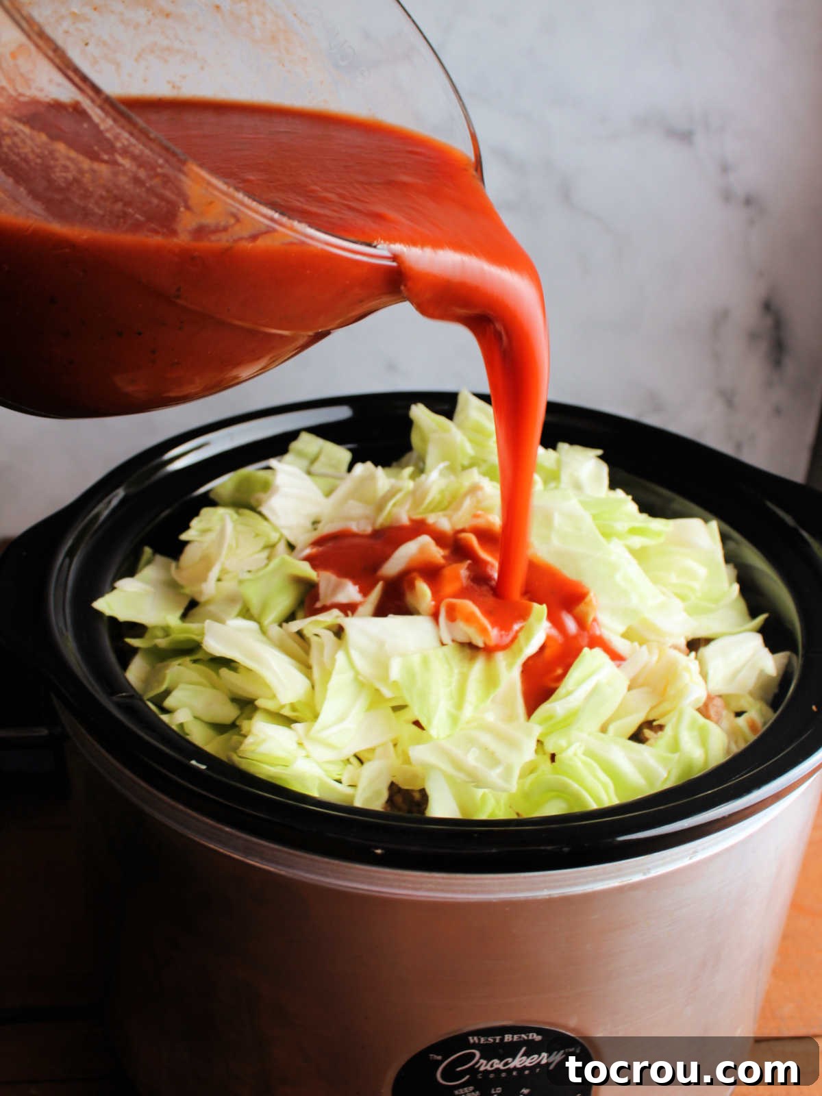 Pouring tomato sauce mixture over slow cooker filled with layers of cabbage, meat, and rice.