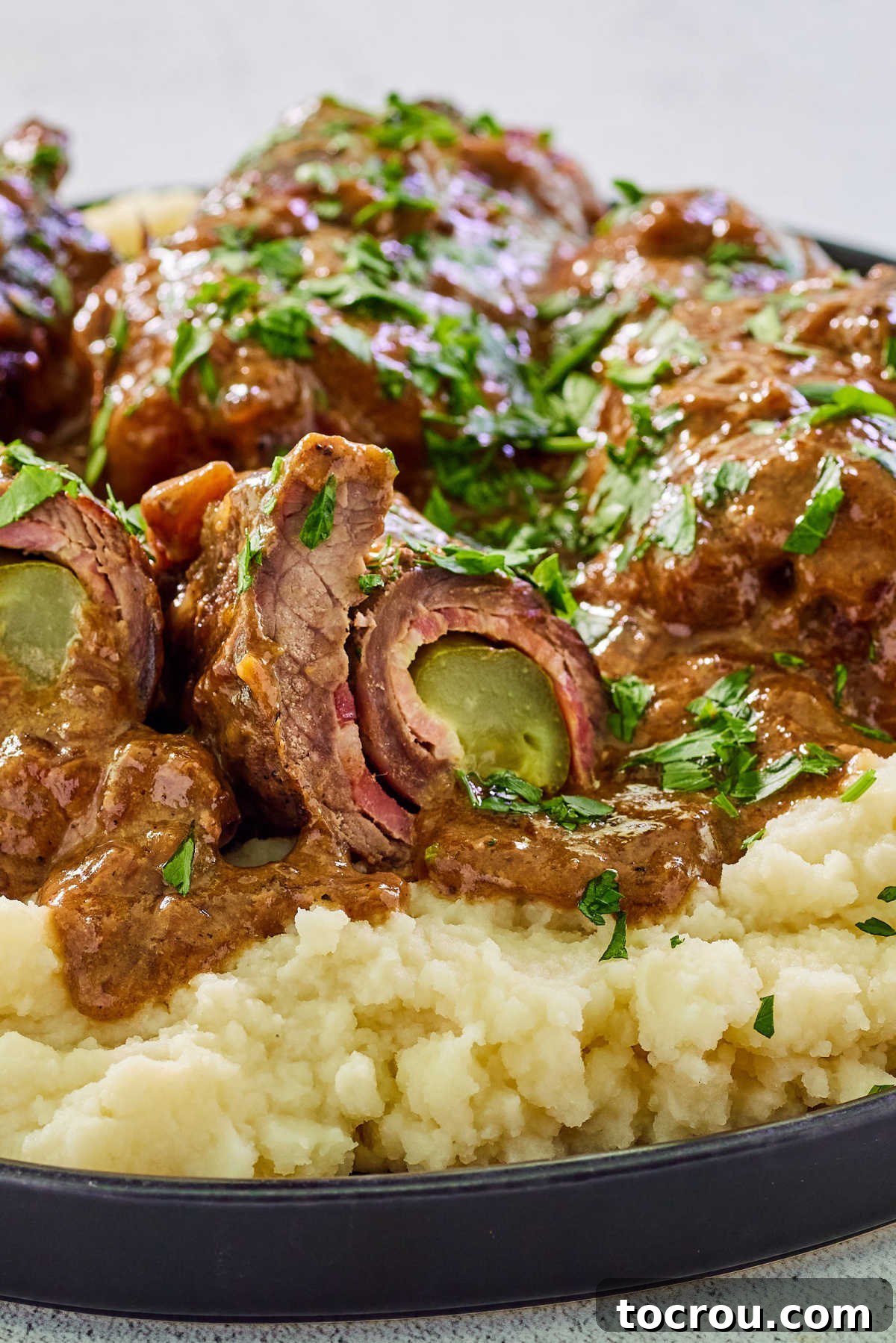 Inside of a cooked rouladen showing tender beef wrapped around a pickle spear and bacon with rich gravy on the outside. 