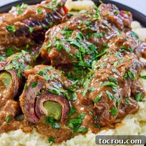 German beef rouladen in rich brown gravy over mashed potatoes with one cut open showing the pickle, onions, and bacon inside.