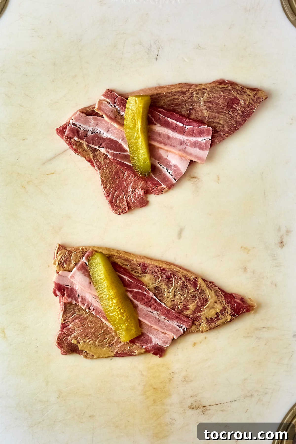 Two thin beef steaks smeared with mustard topped with a strip of bacon and a pickle spear. 