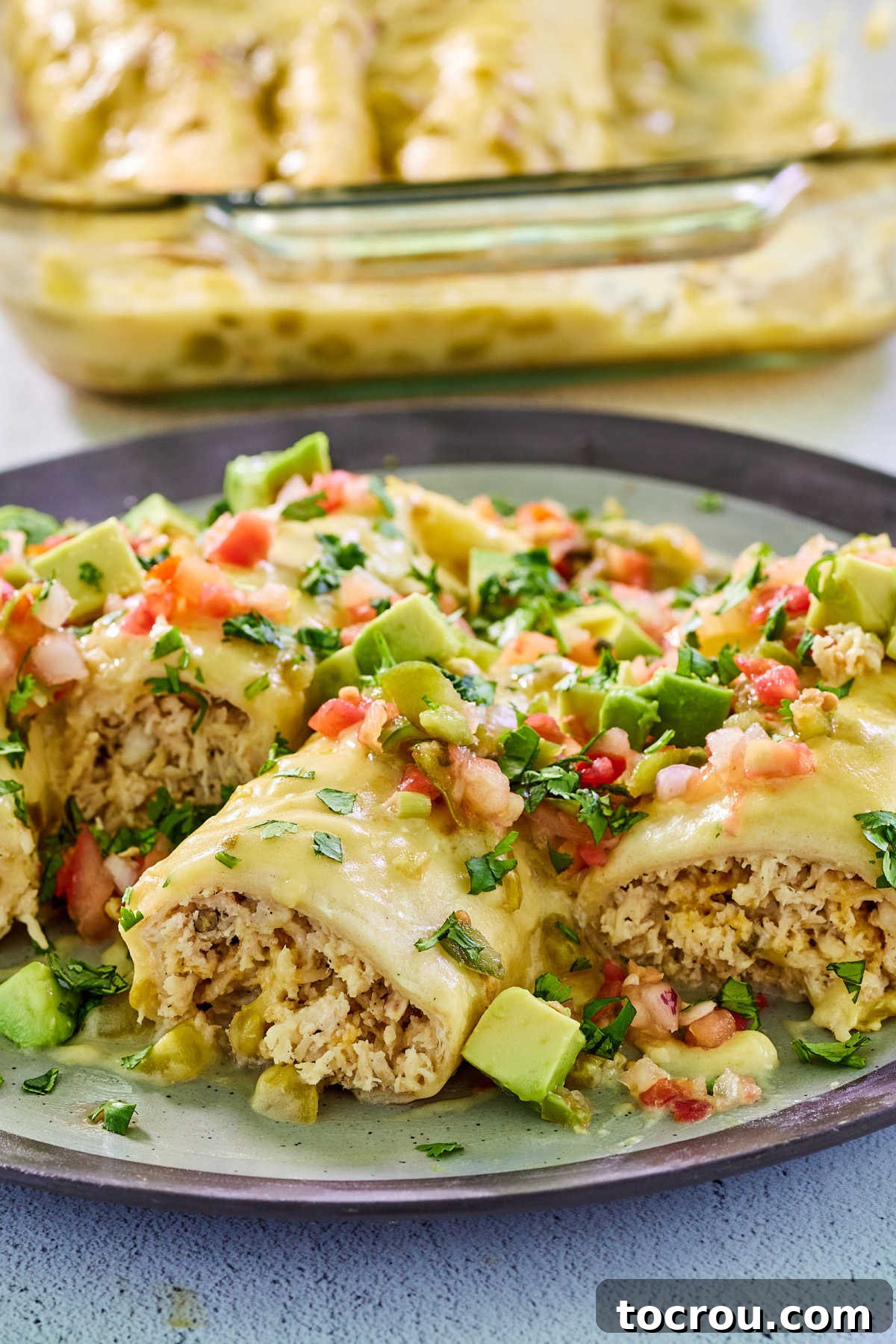 Topped Creamy Chicken Enchiladas Close-up view of a serving of white chicken enchiladas with creamy sauce, showing shredded chicken, and various toppings like avocado, tomatoes, and cilantro, ready to be enjoyed.