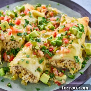 Close dinner plate with several chicken enchiladas with creamy white cheese sauce, topped with avocadoes, tomatoes, and cilantro.