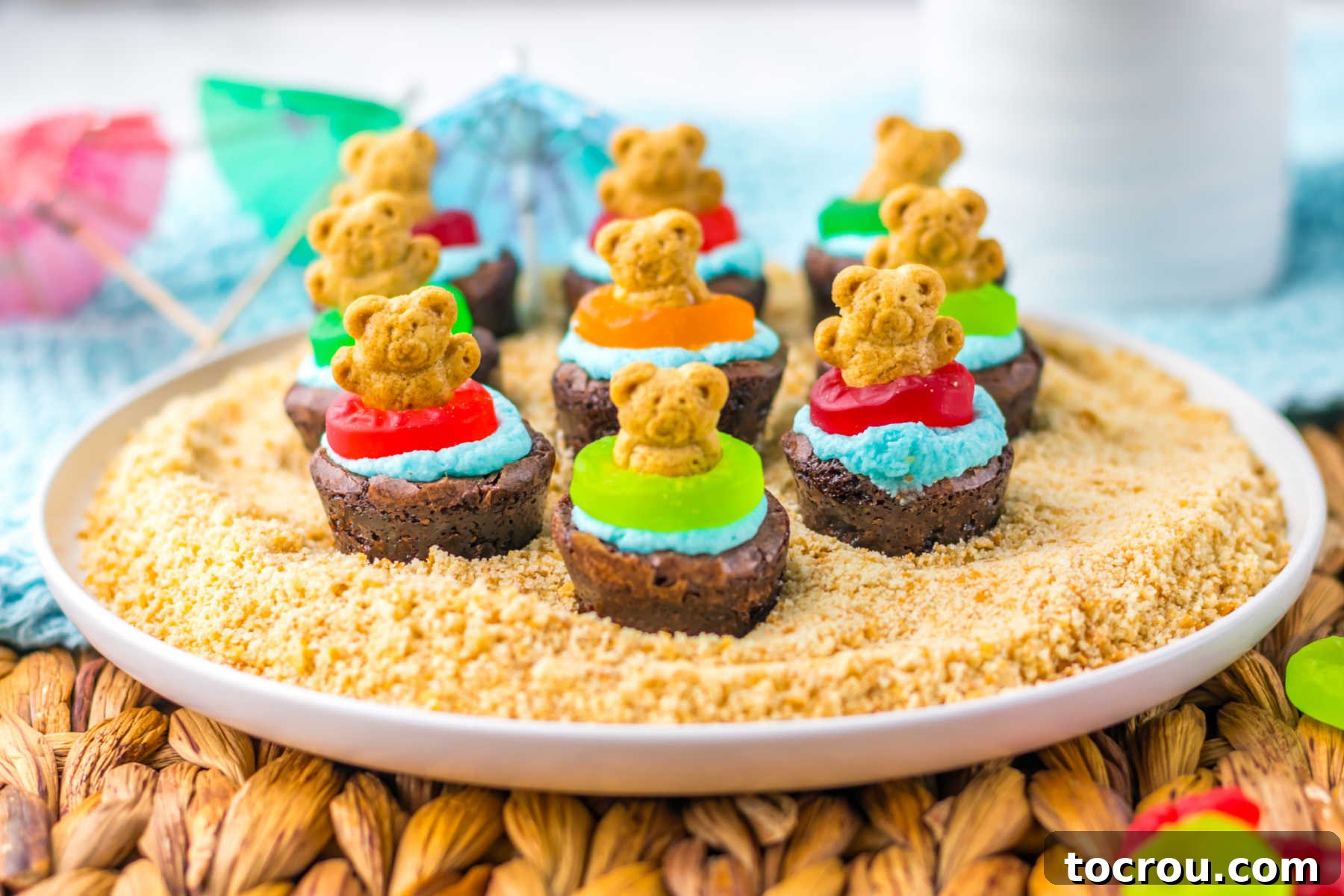 Beach Brownie Bites with Teddy Grahams Mini brownies topped with blue buttercream for water, gummy lifesaver floats and teddy graham bears on a plate of graham cracker crumb sand.