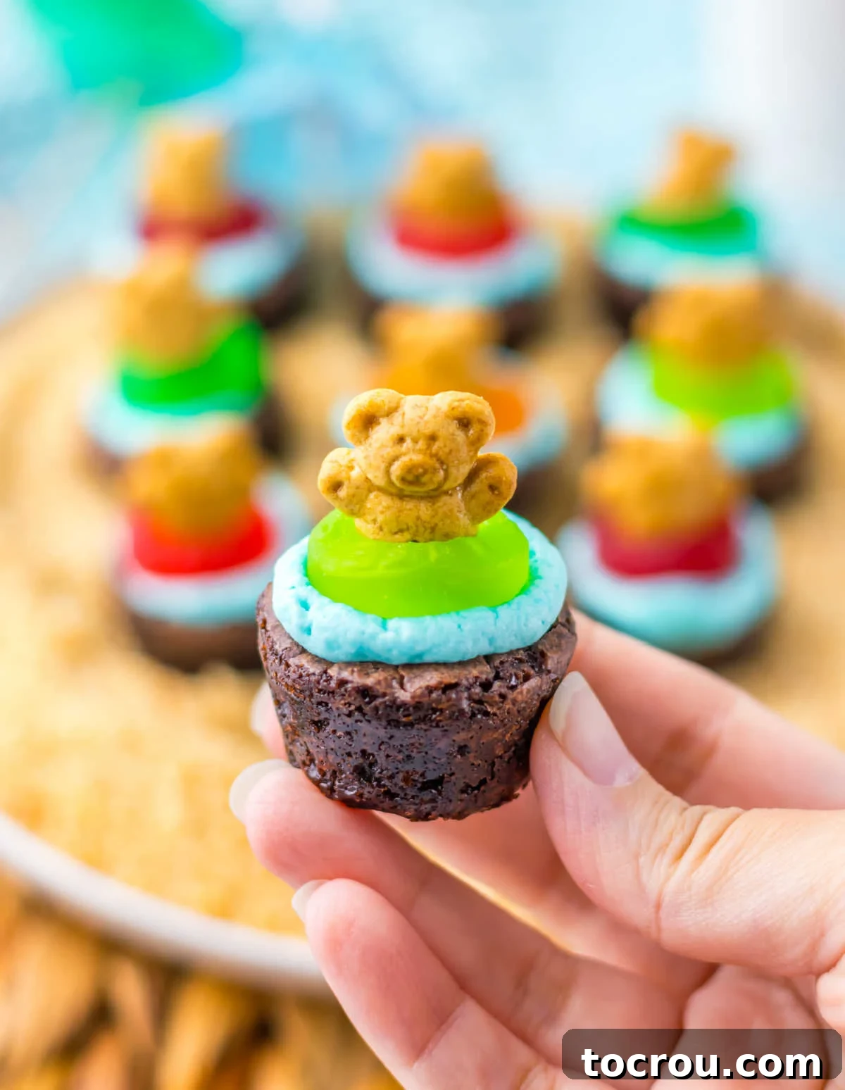 Close-up of Beach Brownie Bite Hand holding a brownie bite topped with blue frosting water, a green gummy life preserver, and a teddy graham, ready to eat.