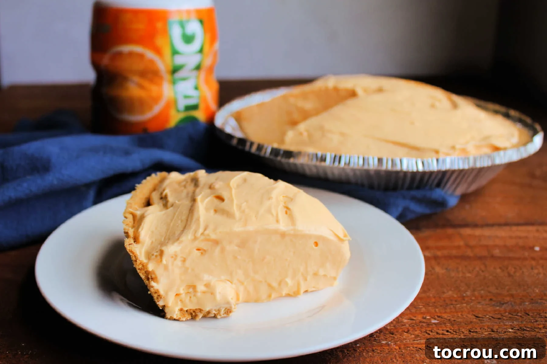 Golden Tang Delicacy 2 Slice of no bake Tang pie with smooth creamy texture on plate with container of Tang powder and pie in background.