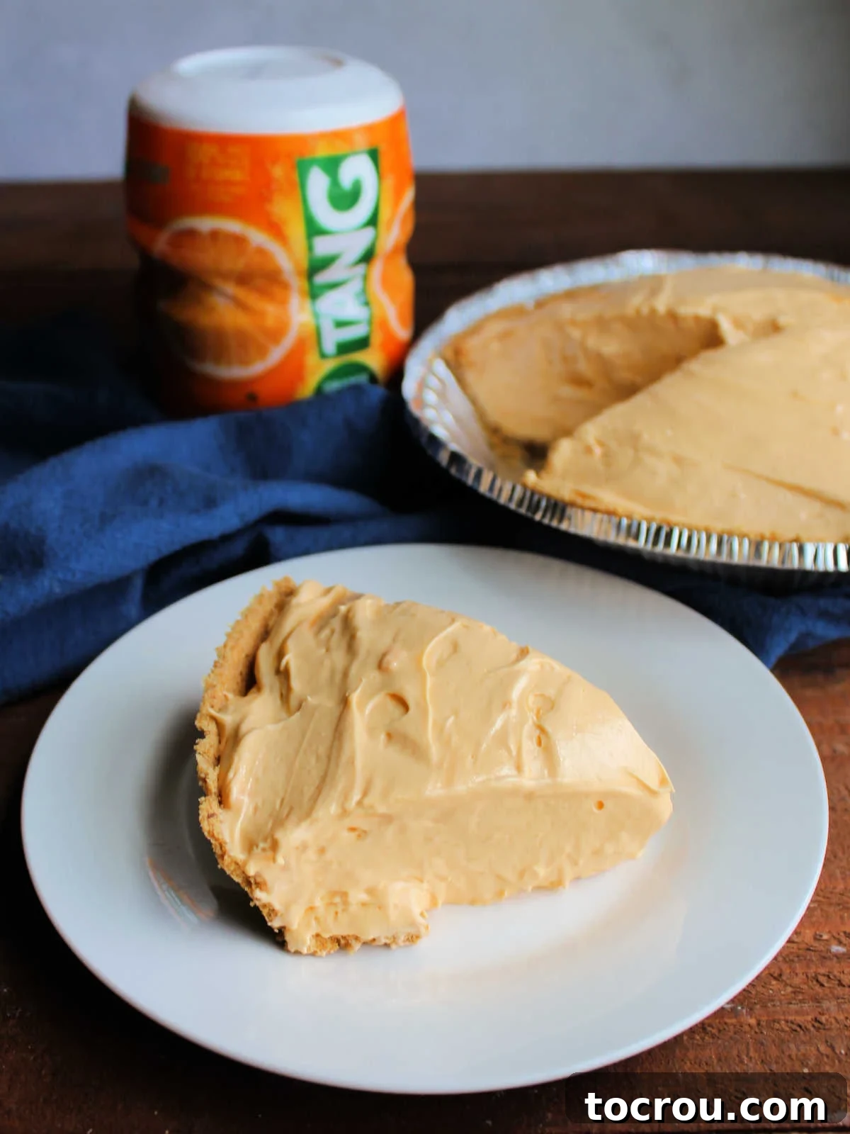 Golden Tang Delicacy 8 Slice of tang pie with smooth light orange filling in graham cracker crust on plate with container of Tang powder and pie in the background.
