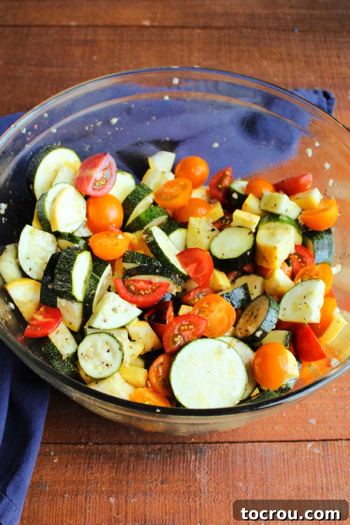 Mixing bowl with sliced zucchini, yellow squash, and halved cherry tomatoes, thoroughly tossed in olive oil with oregano, basil, salt, and pepper.