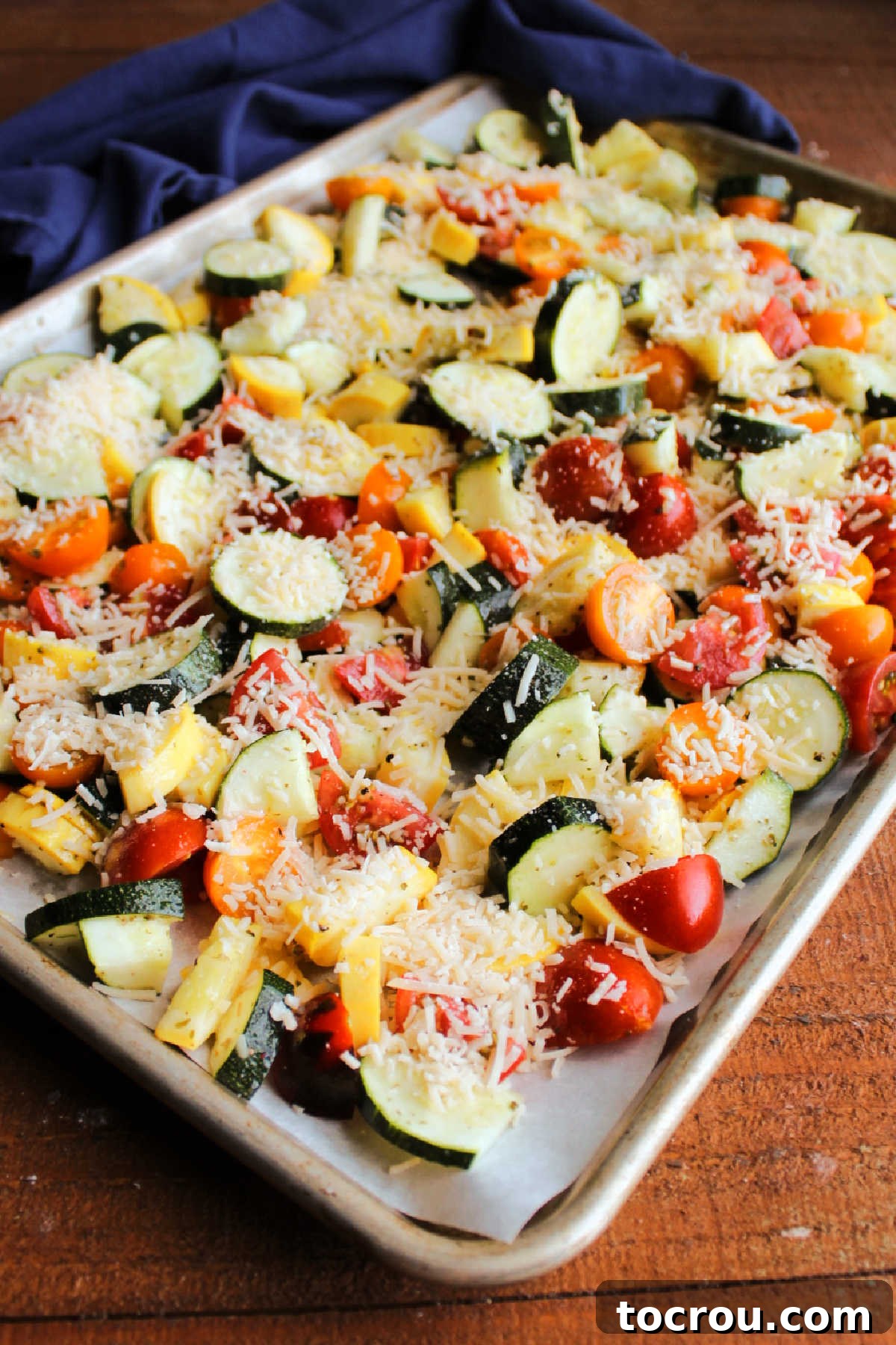 Raw sliced zucchini, yellow squash, and cherry tomatoes evenly spread on a parchment-lined sheet pan, generously topped with grated Parmesan cheese, ready for the oven.