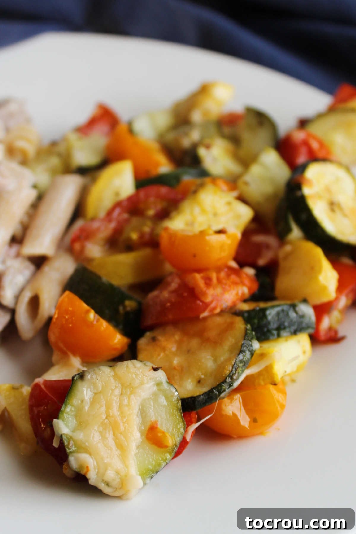 A plate of baked zucchini and tomatoes with melted cheese, served alongside a portion of pasta, ready for a delicious meal.