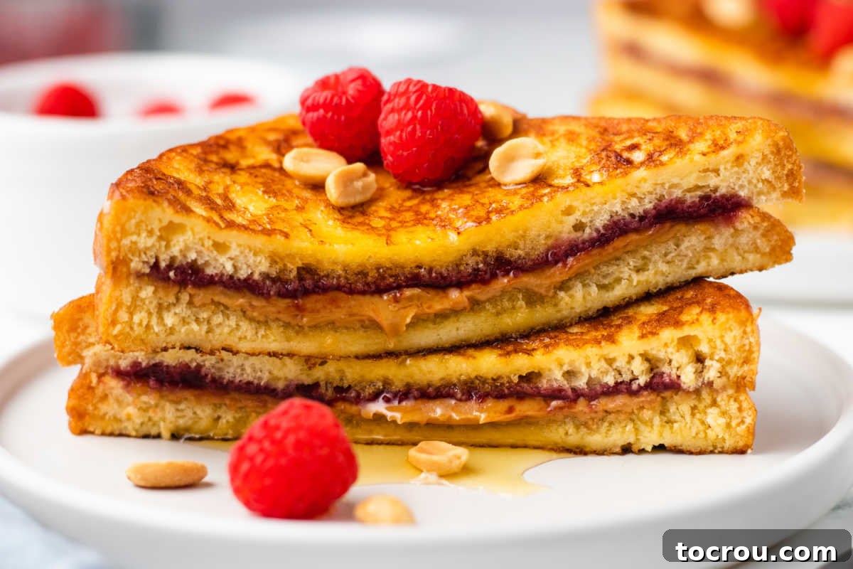 Peanut Butter and Jelly Stuffed French Toast 2 Two peanut butter and jelly french toast halves stacked on each other showing the gooey filling and browned exterior served with fresh raspberries and peanuts.