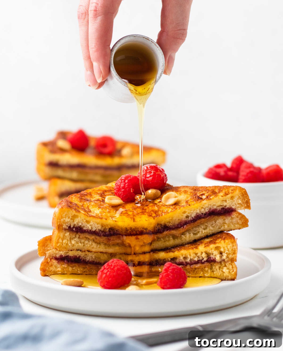 Peanut Butter and Jelly Stuffed French Toast 8 Hand pouring a small container of maple syrup over a plate of peanut butter and jelly french toast, ready to eat.