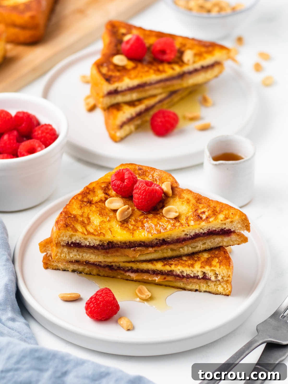 Peanut Butter and Jelly Stuffed French Toast 9 Two plates of prepared peanut butter and jelly french toast garnished with raspberries, peanuts, and maple syrup, ready to eat.