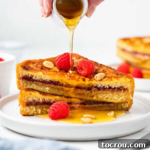 Peanut Butter and Jelly Stuffed French Toast 10 Pouring maple syrup over a stack of peanut butter and jelly stuffed french toast served with fresh raspberries and peanuts.