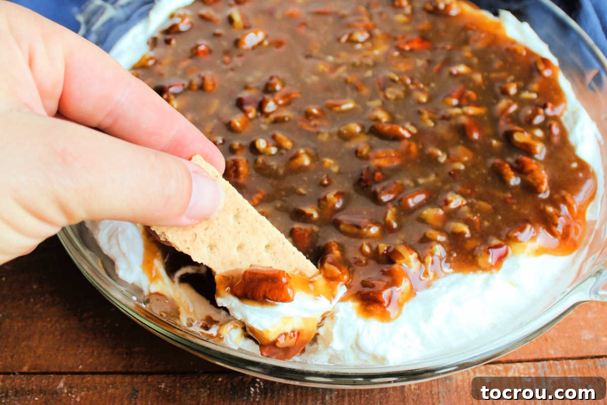 Hand dipping a piece of graham cracker into pecan pie dip getting some cream cheese mixture and some brown sugar pecan mixture on the graham cracker.