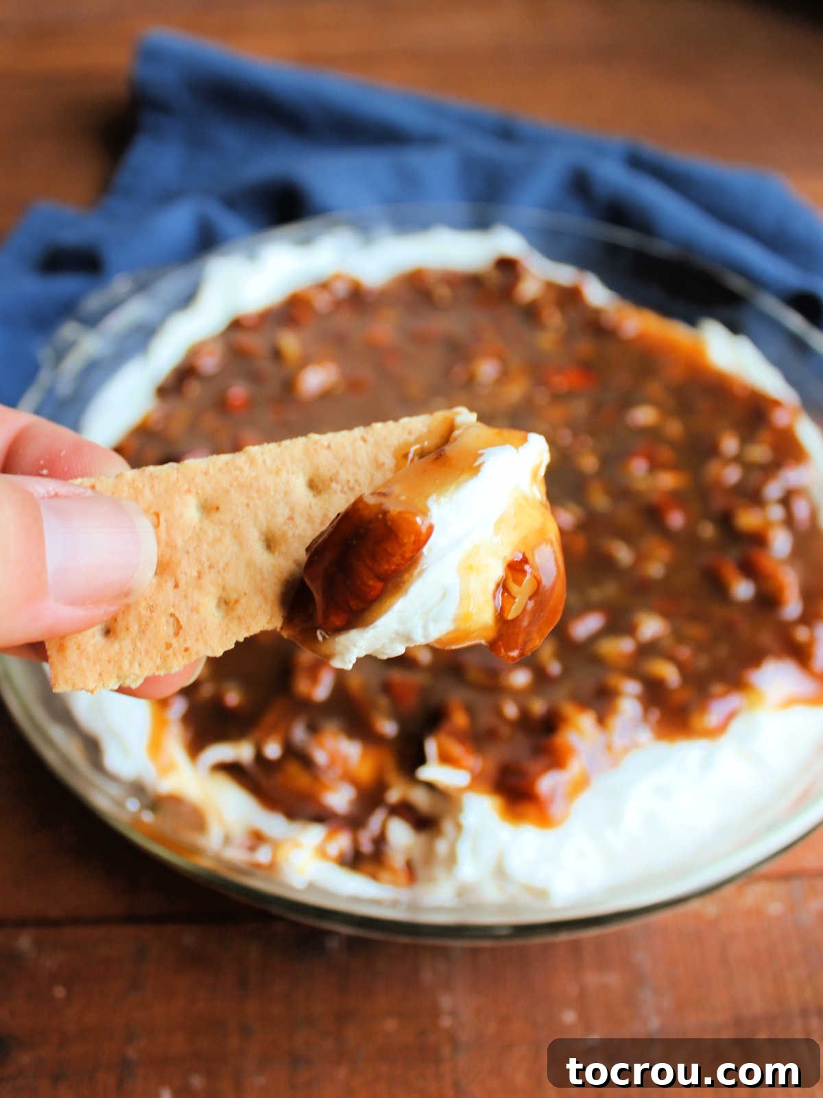 Hand holding graham cracker with pecan pie dip on it showing creamy base and gooey pecan layer.
