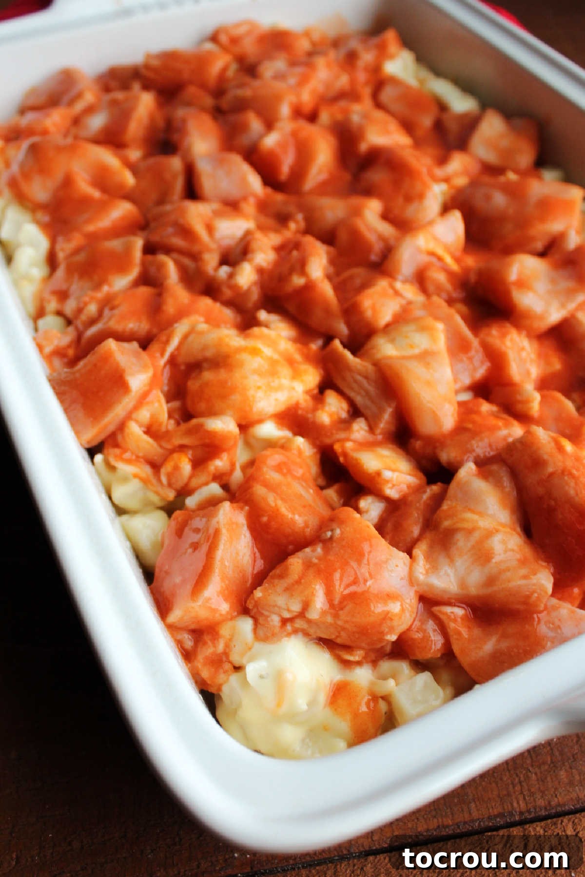 Raw chicken chunks generously coated in vibrant buffalo sauce, arranged neatly over the cheesy ranch potato layer in a casserole pan before baking.