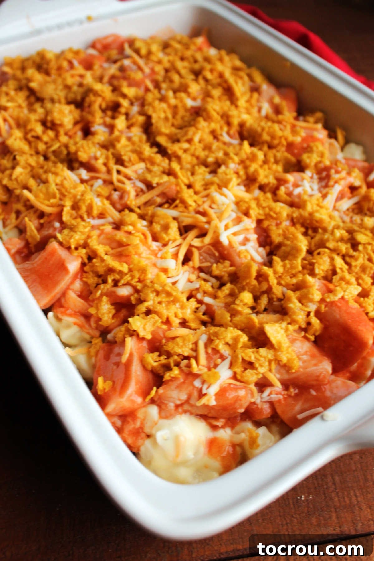 Unbaked casserole showing buffalo chicken and cheesy ranch potatoes topped with an even layer of shredded cheese and golden, buttered corn flakes, ready for the oven.