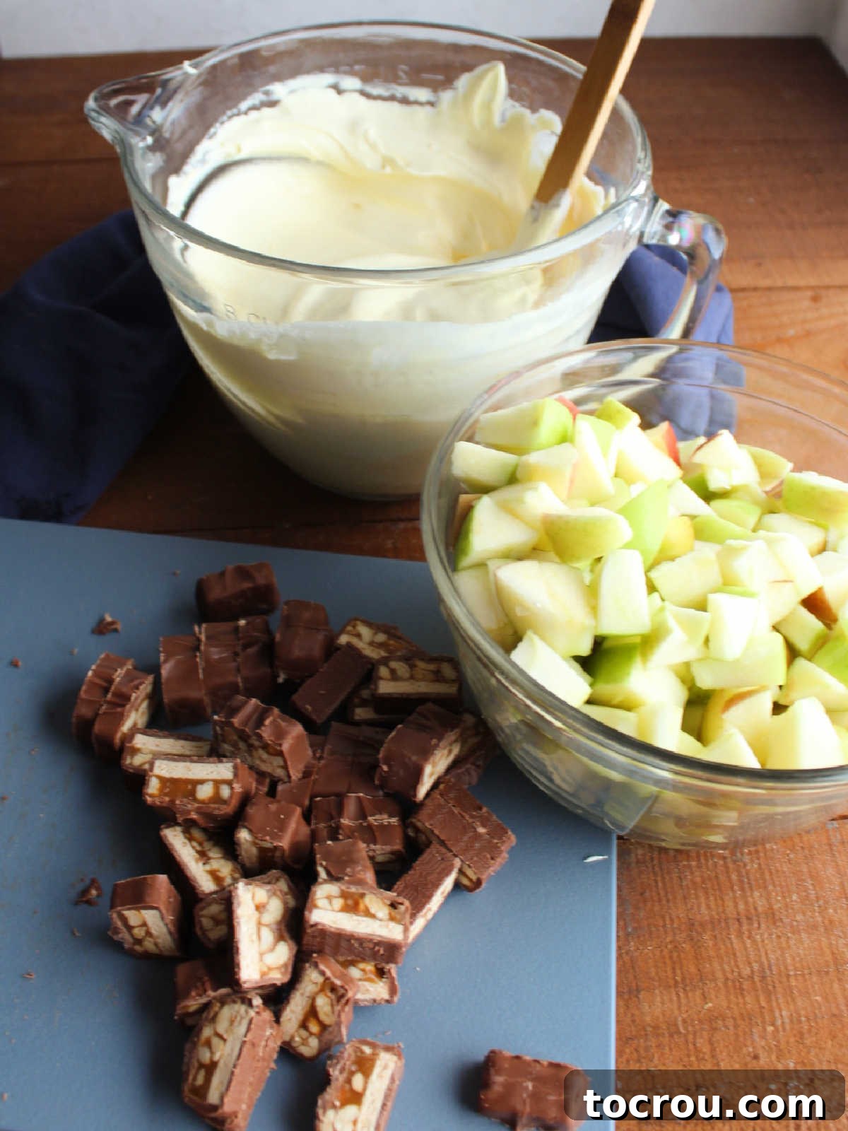 Decadent Apple Snickers Dessert 4 Bowl of chopped apples tossed in lemon juice next to chunks of snickers on a cutting board and a bowl of vanilla pudding and whipped topping mixture.