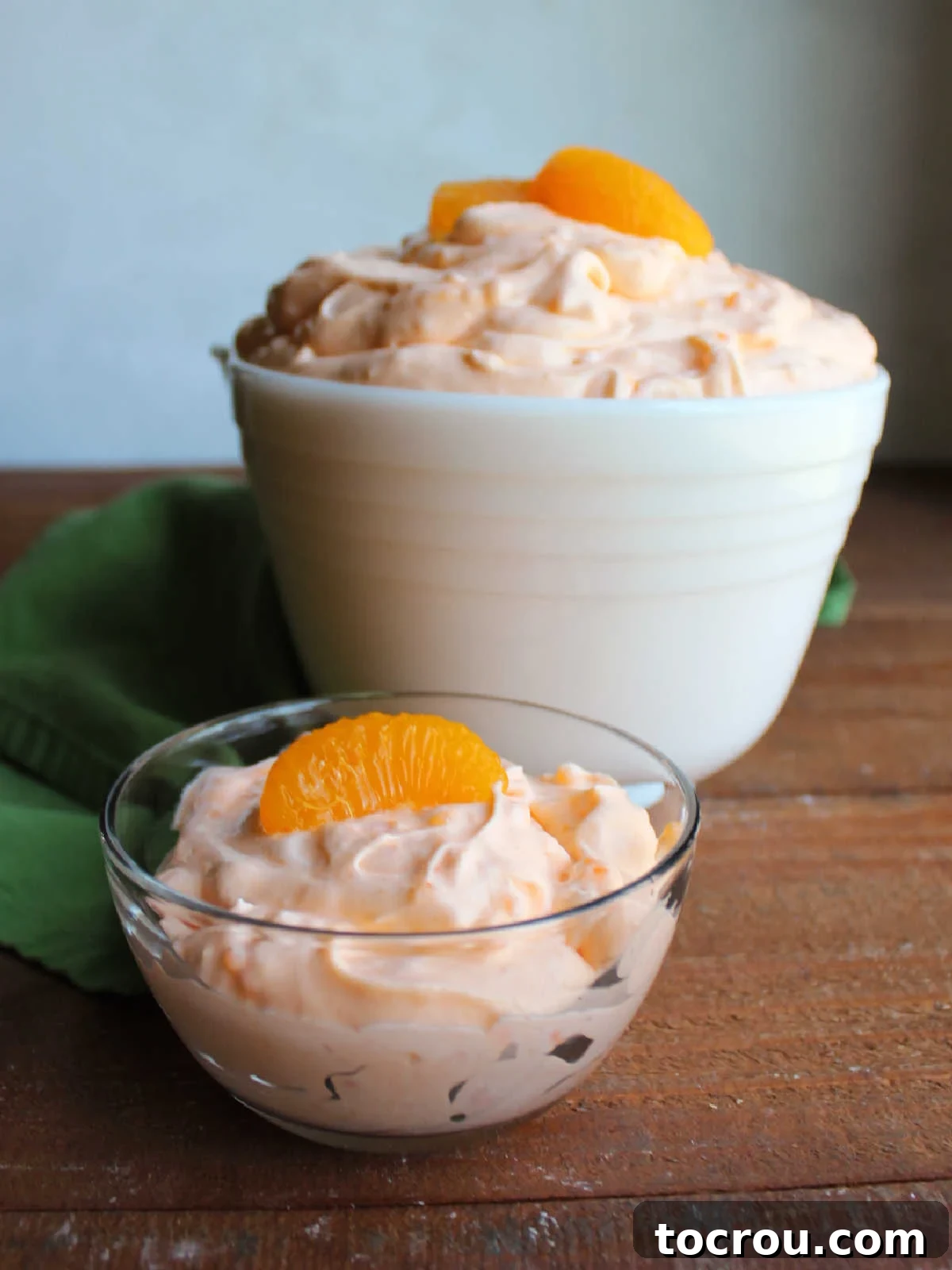Small glass bowl with a helping of tapioca orange fluff inside with a large serving bowl of the mixture in the background. 
