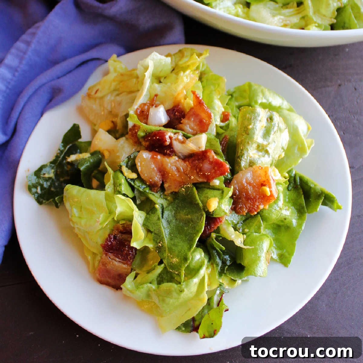 A small plate generously piled high with wilted lettuce salad, featuring big chunks of crispy bacon and slices of hard-boiled egg mixed throughout.